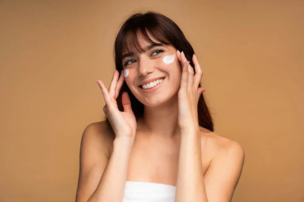 A smiling woman applying cream cleanser to her face with her fingers, showcasing a skincare routine.