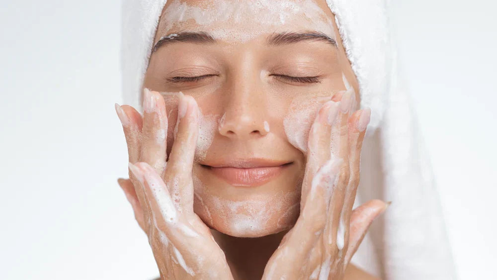 A woman with a towel wrapped around her head, gently massaging foam cleanser onto her face, smiling with her eyes closed.