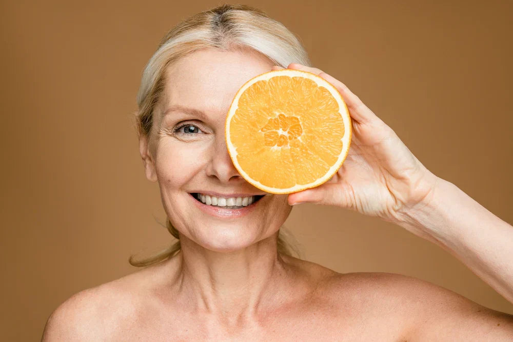 A smiling woman holding a slice of orange up to her eye, showcasing a fresh, healthy glow, representing vitamin C benefits for skin.