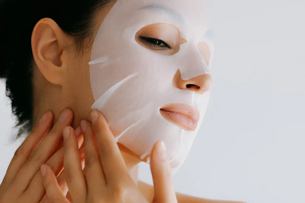 A woman applying a sheet face mask to her face, gently pressing it against her skin with a relaxed expression.