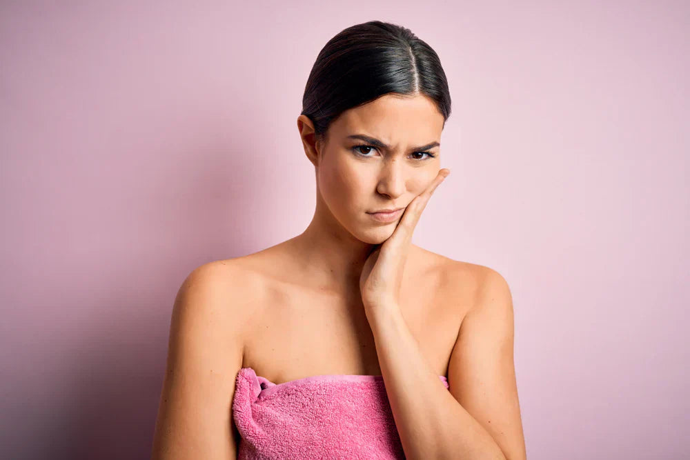 A woman with a towel wrapped around her, standing against a pink background, looking concerned and touching her cheek, representing skin dullness.