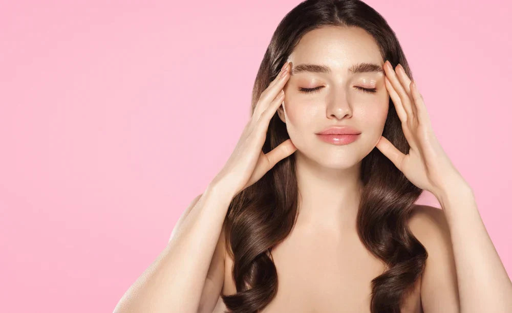 A youthful woman gently massaging her temples, smiling with her eyes closed, against a soft pink background, suggesting relaxation and healthy collagen levels.