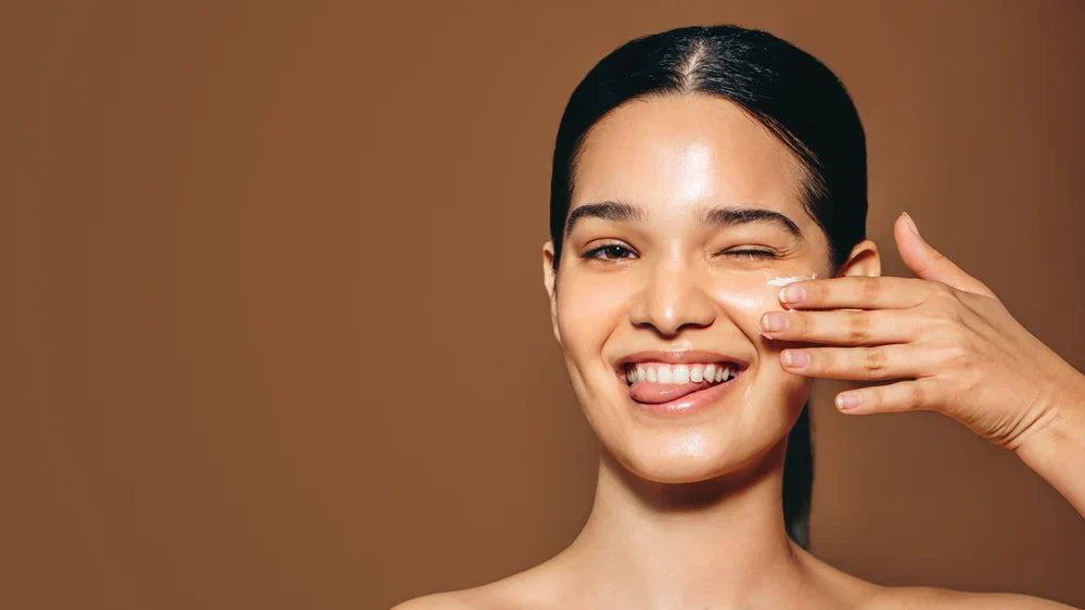 A woman smiling and applying moisturiser to her face, winking playfully while showcasing glowing, hydrated skin.