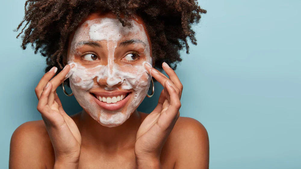 A woman smiling while applying cleanser to her face, with foam visible on her skin, against a blue background.